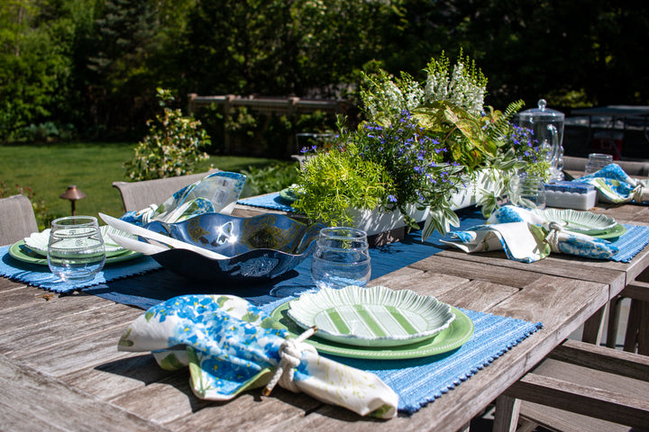 Outdoor table setting with blue and green tablecloth, silverware, and floral centerpieces.